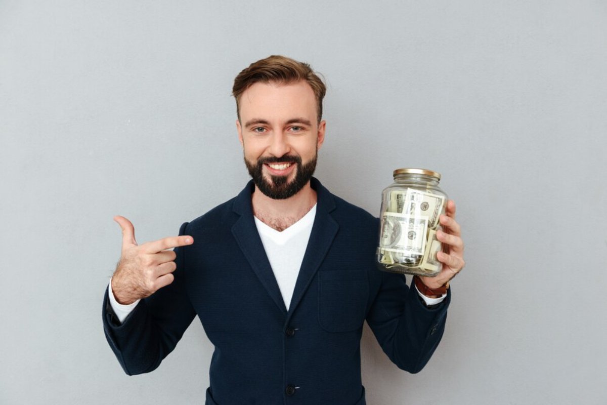 Smiling man in a suit holding a jar of money, pointing at it with his finger.