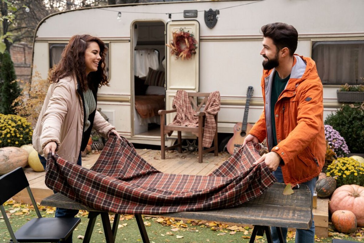 A man and a woman setting a picnic blanket on a table near an RV in autumn.