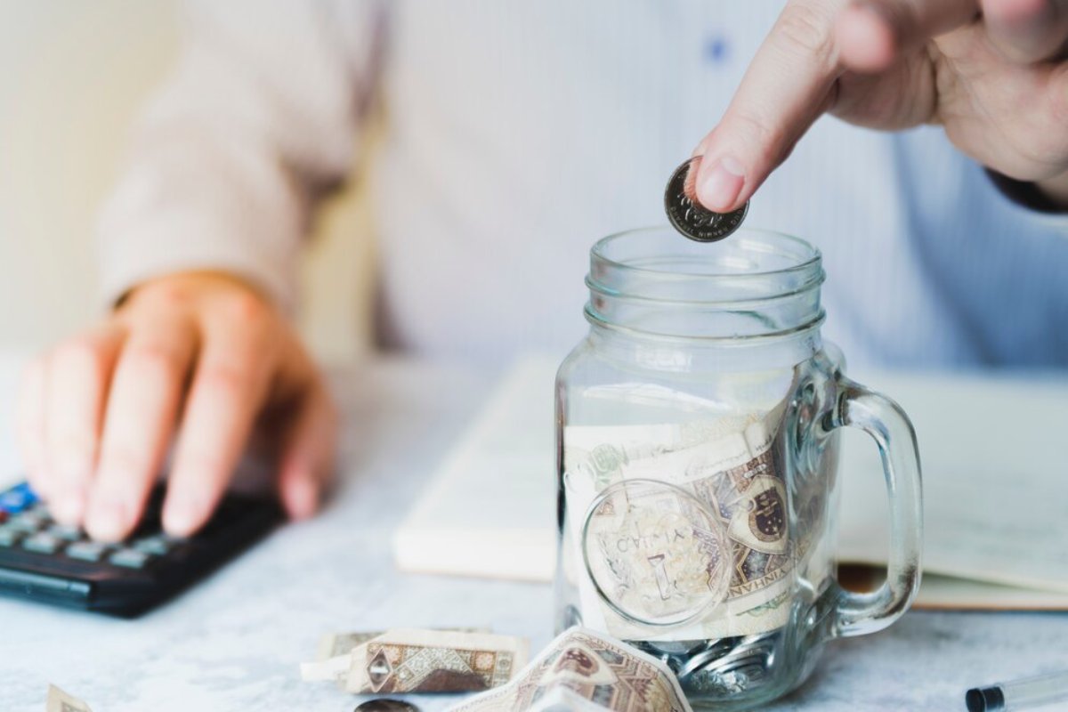 Person sits at a desk, placing a coin into a glass jar filled with money, near calculator and cash.