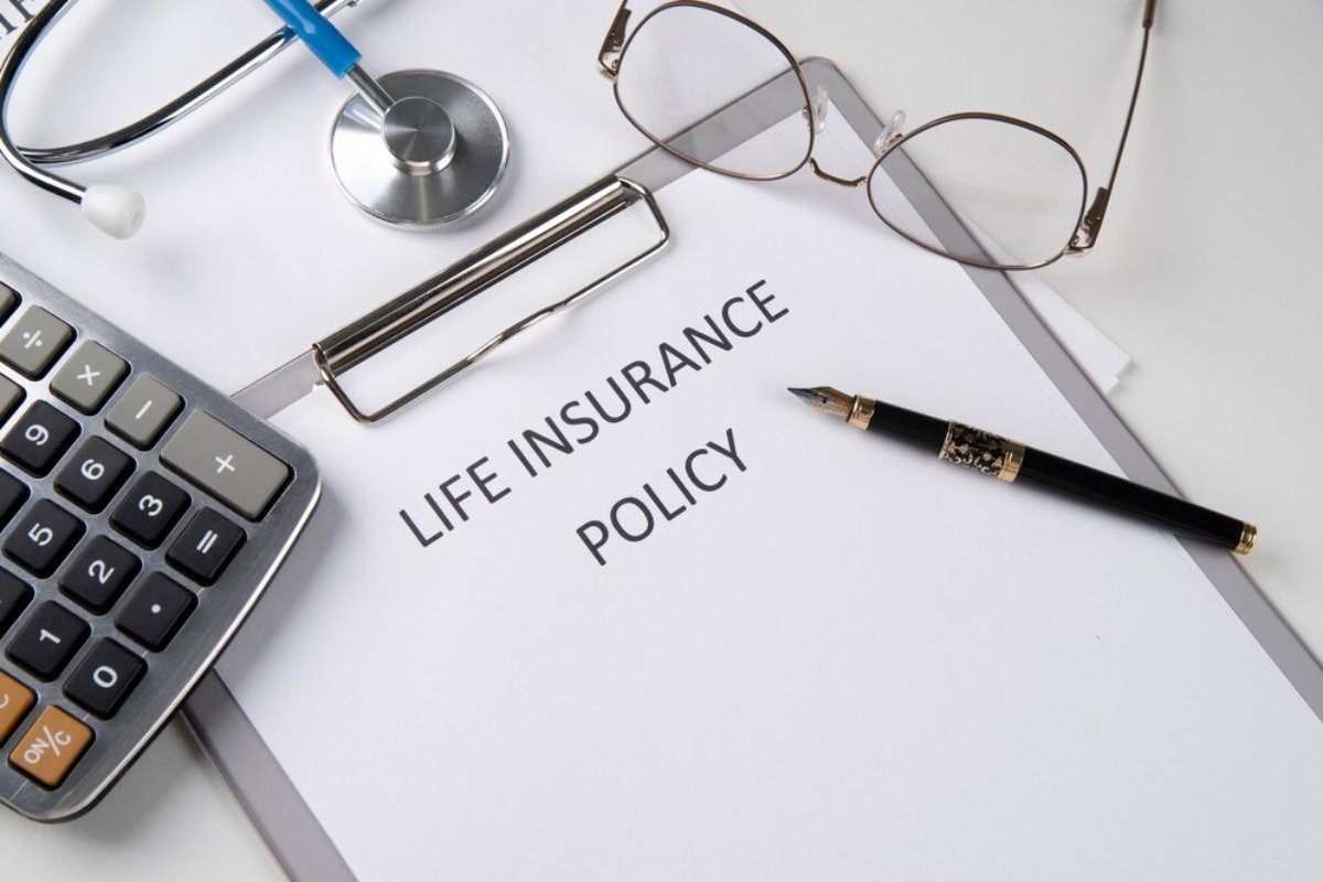 White desk with a life insurance policy document, a calculator, eyeglasses, a pen, and a stethoscope.
