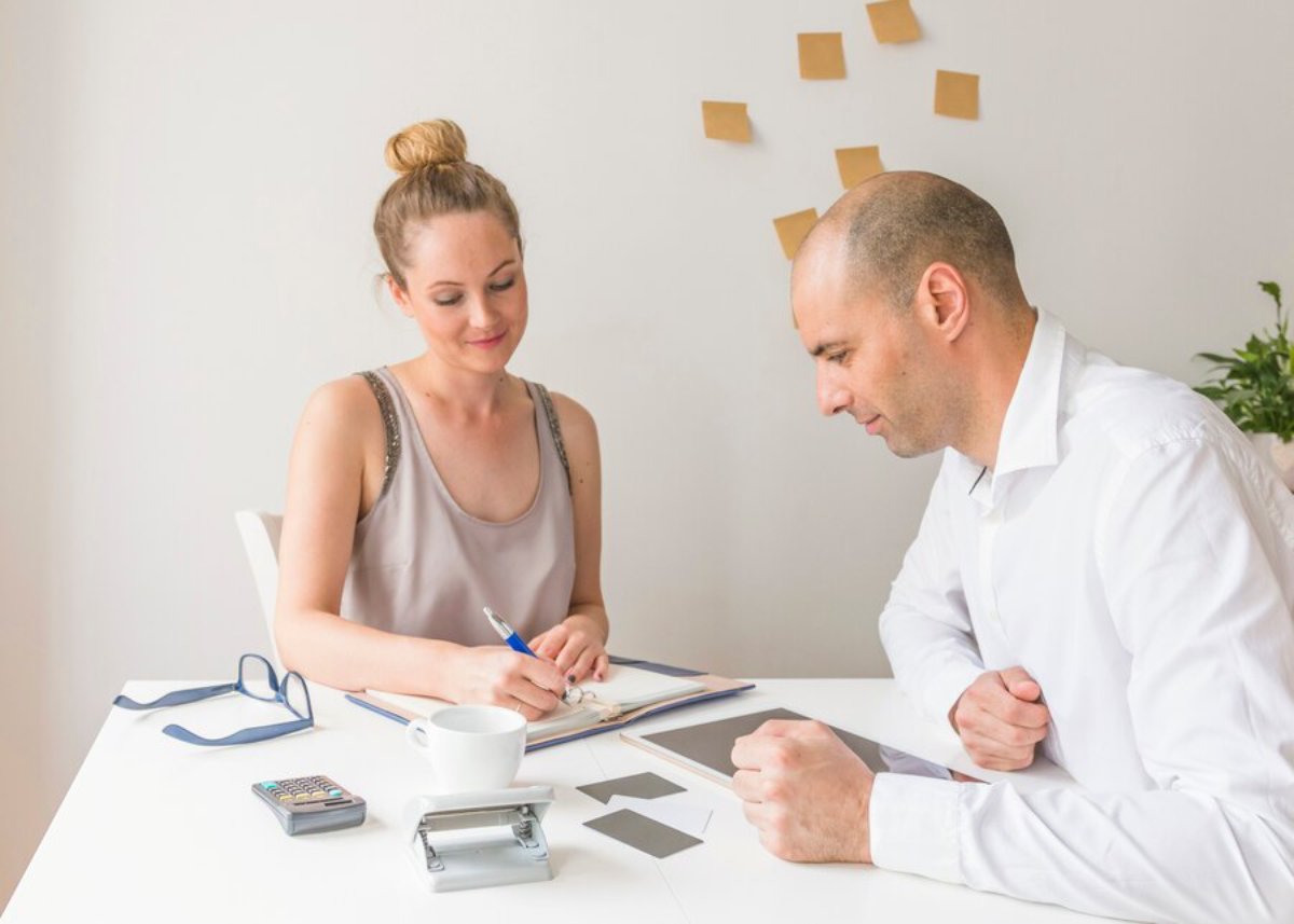 Two professionals, a man and a woman, discussing documents at a white office desk.