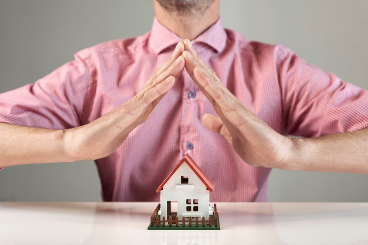 Man in pink shirt forming a roof shape with his hands over a model house on a table.