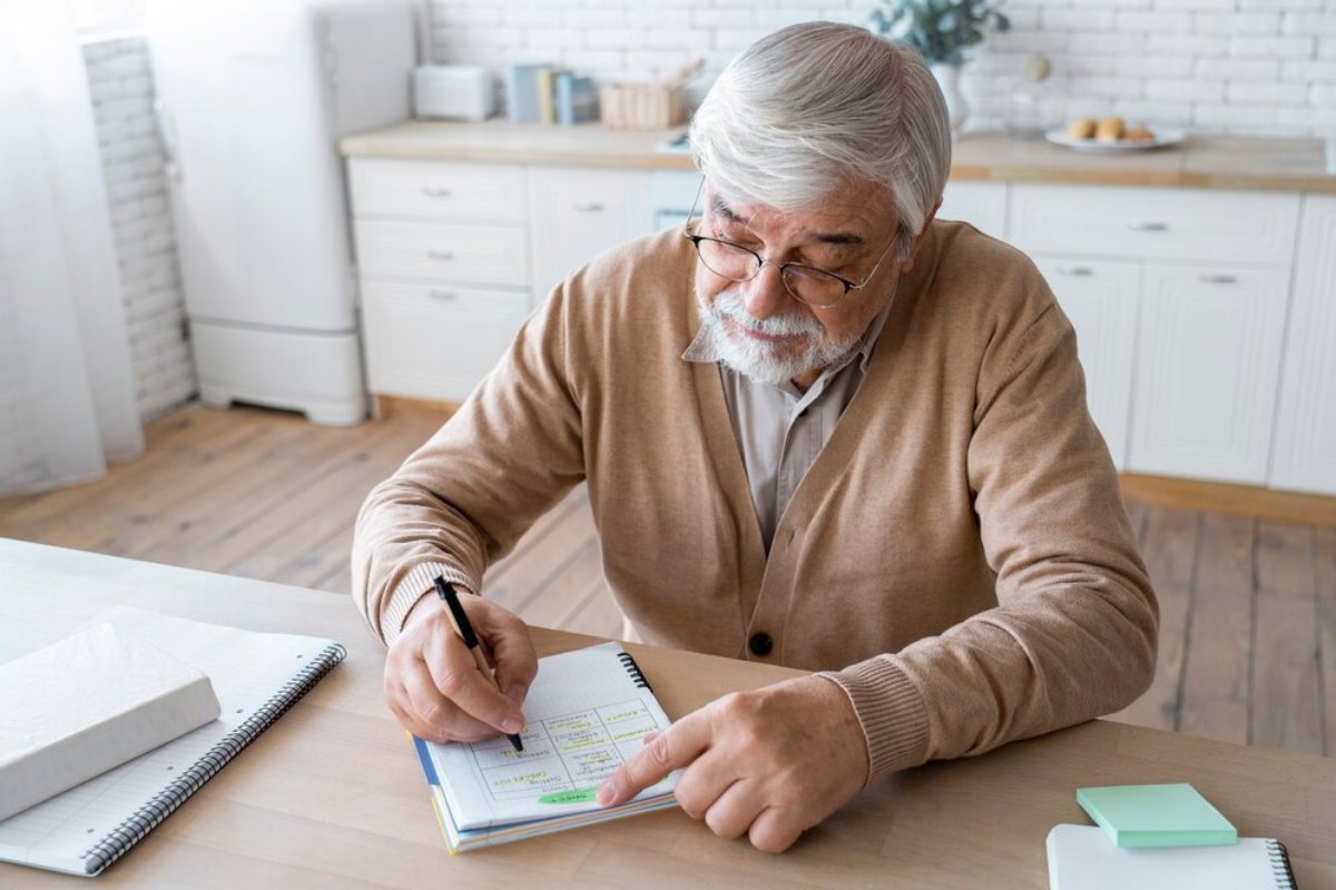 Senior man with glasses writing in a planner at a kitchen table.