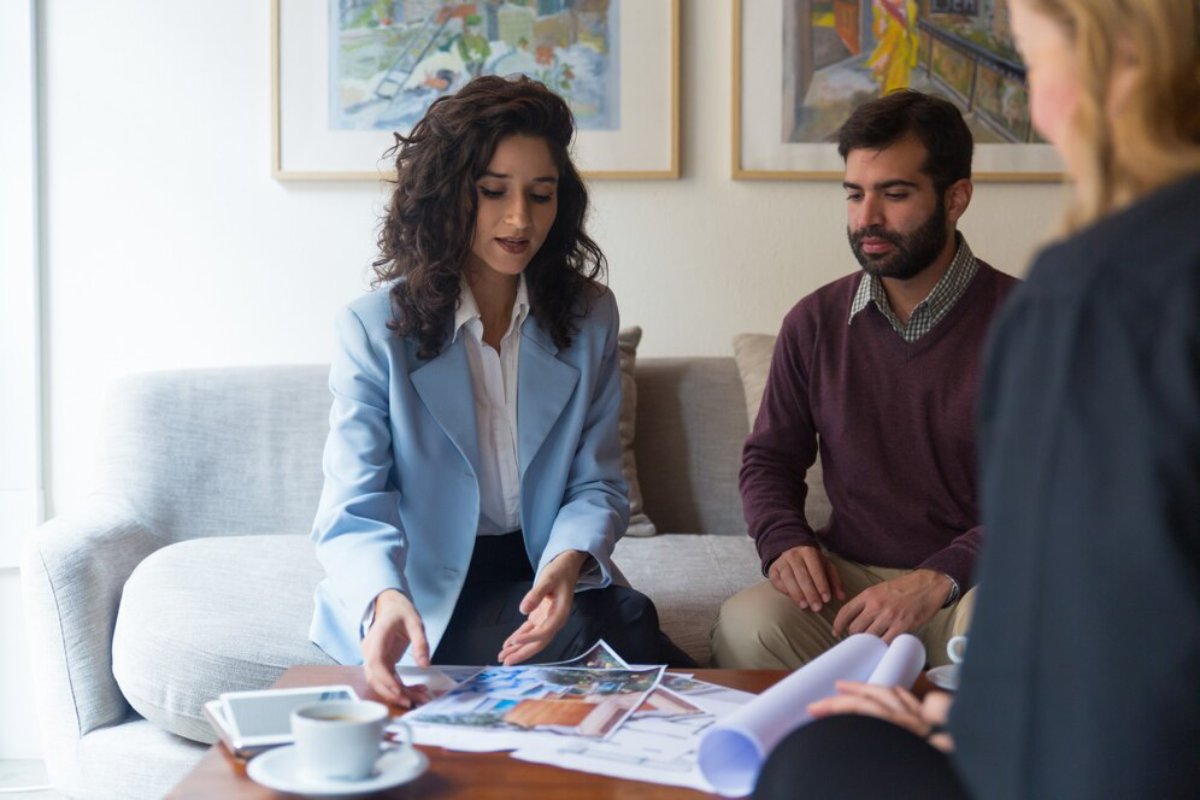 Three professionals sit around a table reviewing documents, with a coffee cup visible, in a brightly lit room.