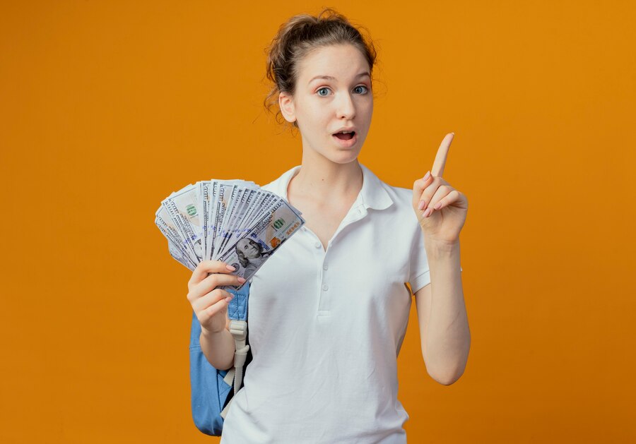Young woman holding cash and pointing upwards, looking surprised, against an orange background.