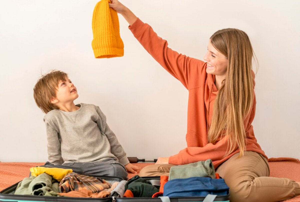 A woman and a young boy smiling while packing a suitcase, the woman holding up a yellow beanie.
