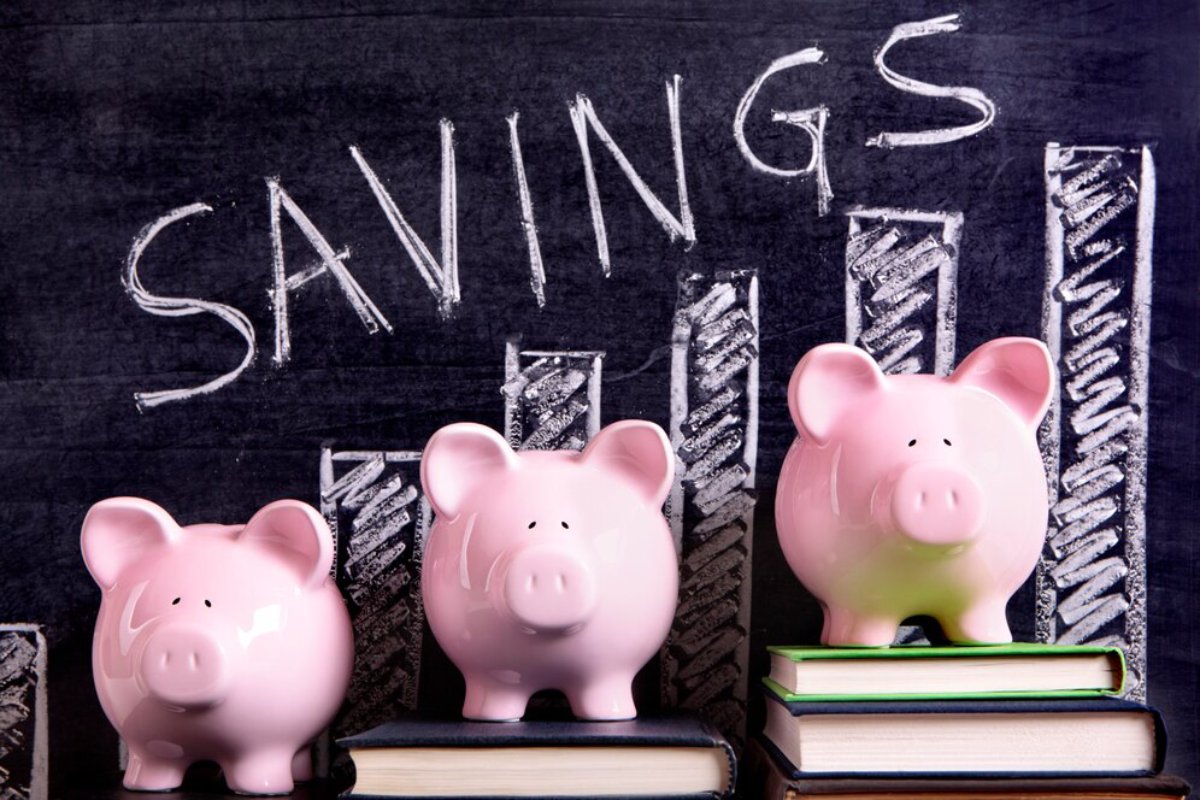 Three pink piggy banks on stacked books against a chalkboard with drawn bar graphs labeled 