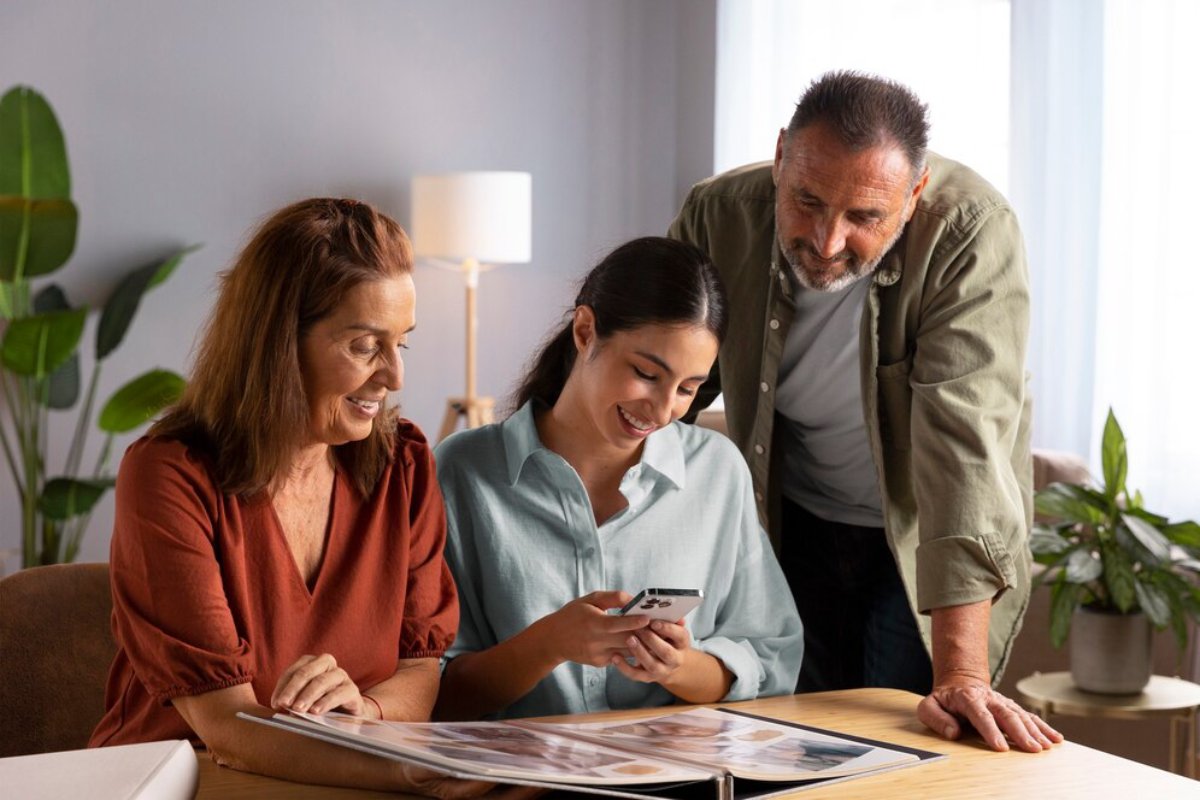 A young woman shows a smartphone to an older couple while looking at photo albums in a cozy living room.