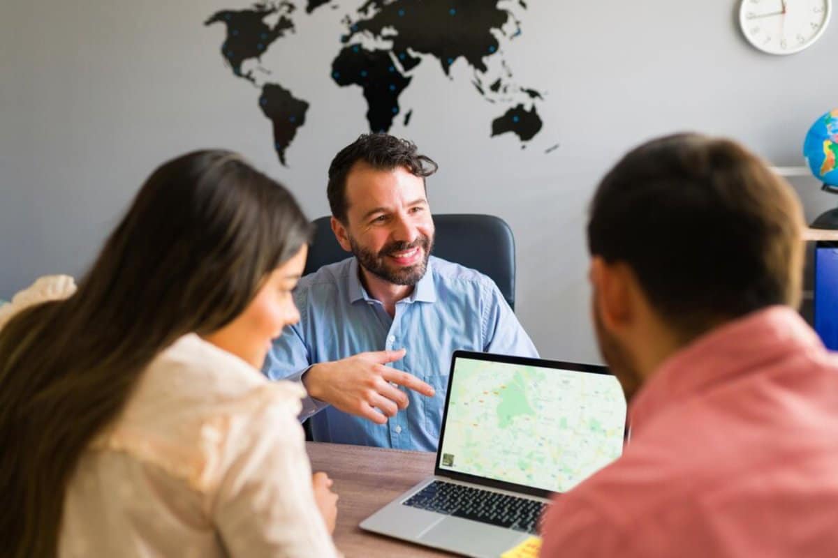 Three people discussing over a laptop with a world map in the background in an office setting.
