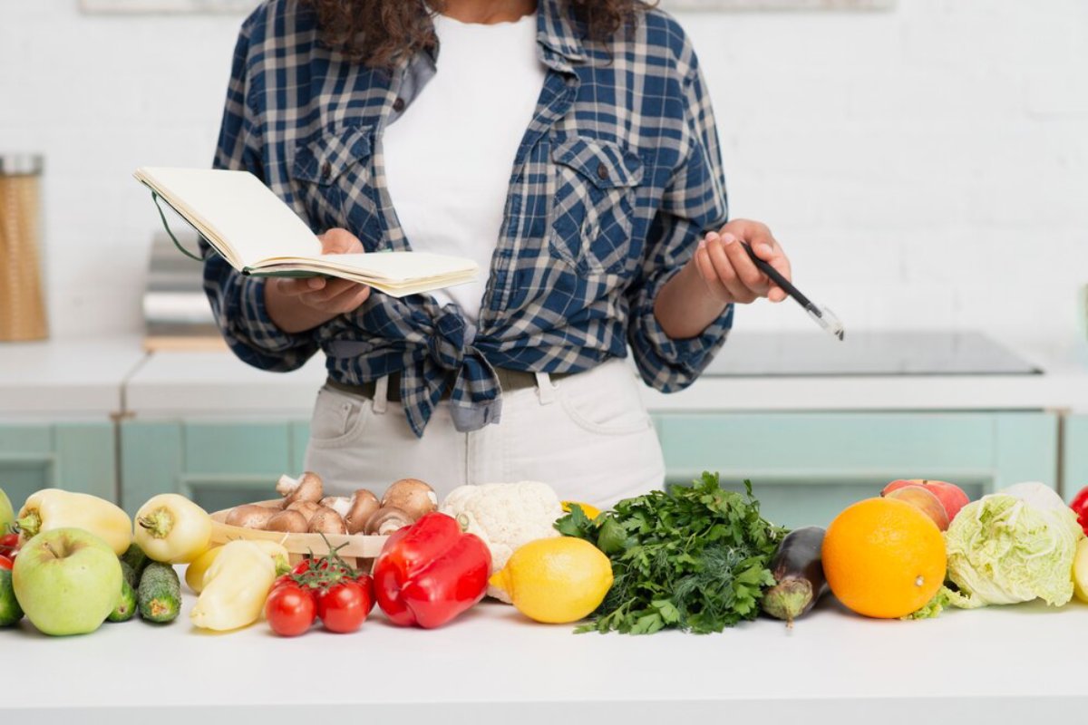 Woman reading a recipe book and holding a spoon, standing behind a counter with various fresh vegetables and fruits.