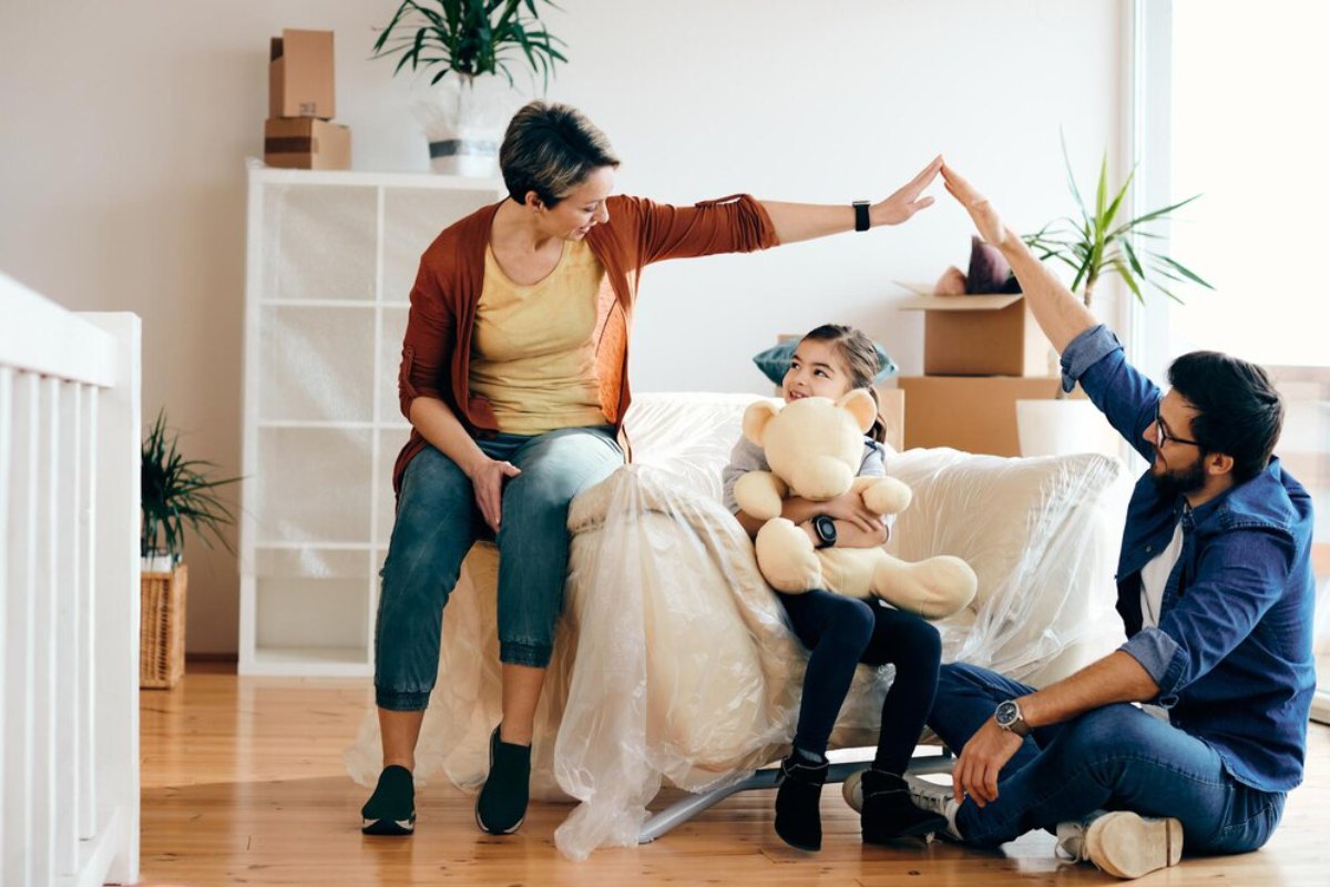A family with a child holding a stuffed toy, smiling and interacting in a living room filled with moving boxes.