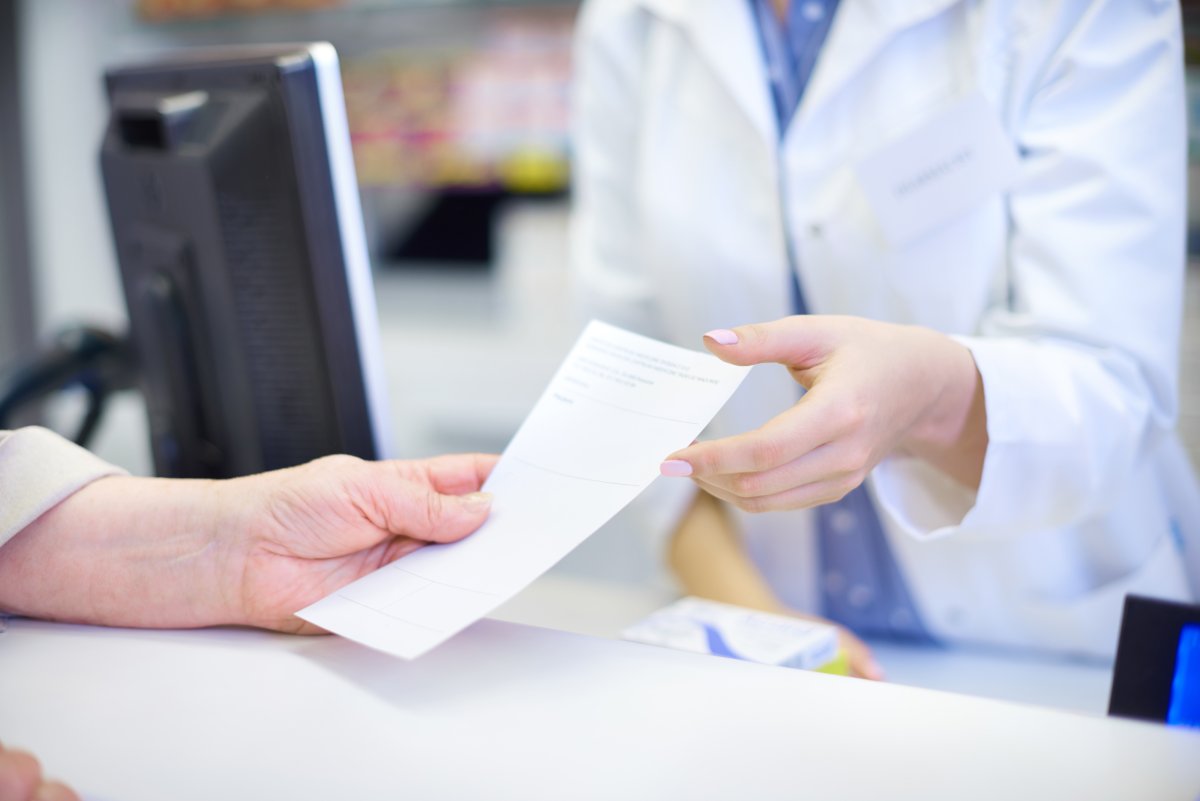 Pharmacist in white coat handing a prescription to a patient across the pharmacy counter.