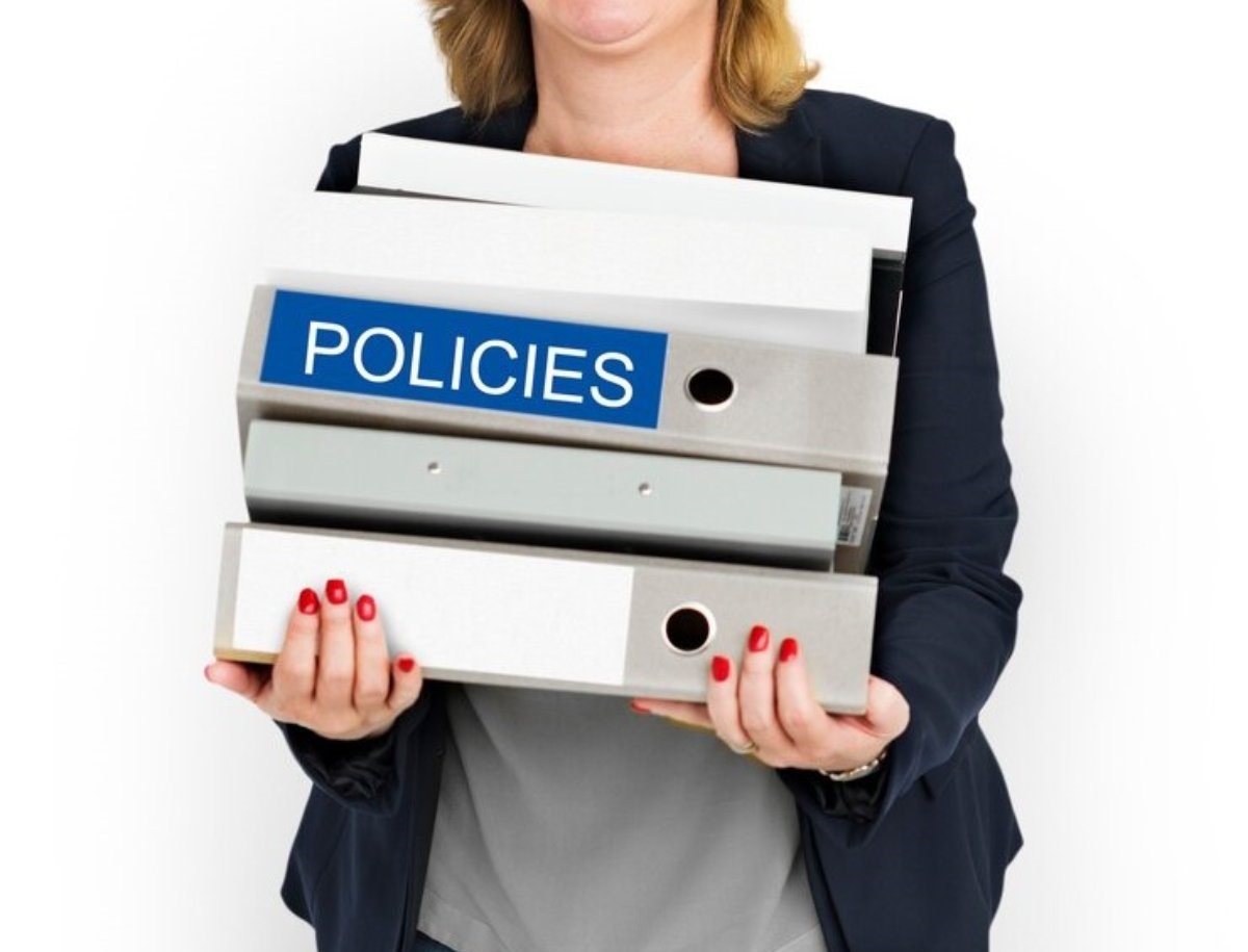 Businesswoman carrying three binders labeled 'POLICIES' against a white background.
