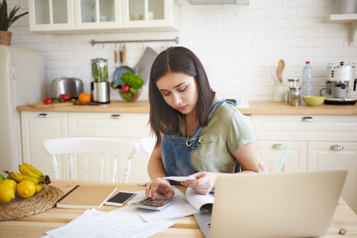 Young woman calculating finances in a bright kitchen with a laptop and paperwork.
