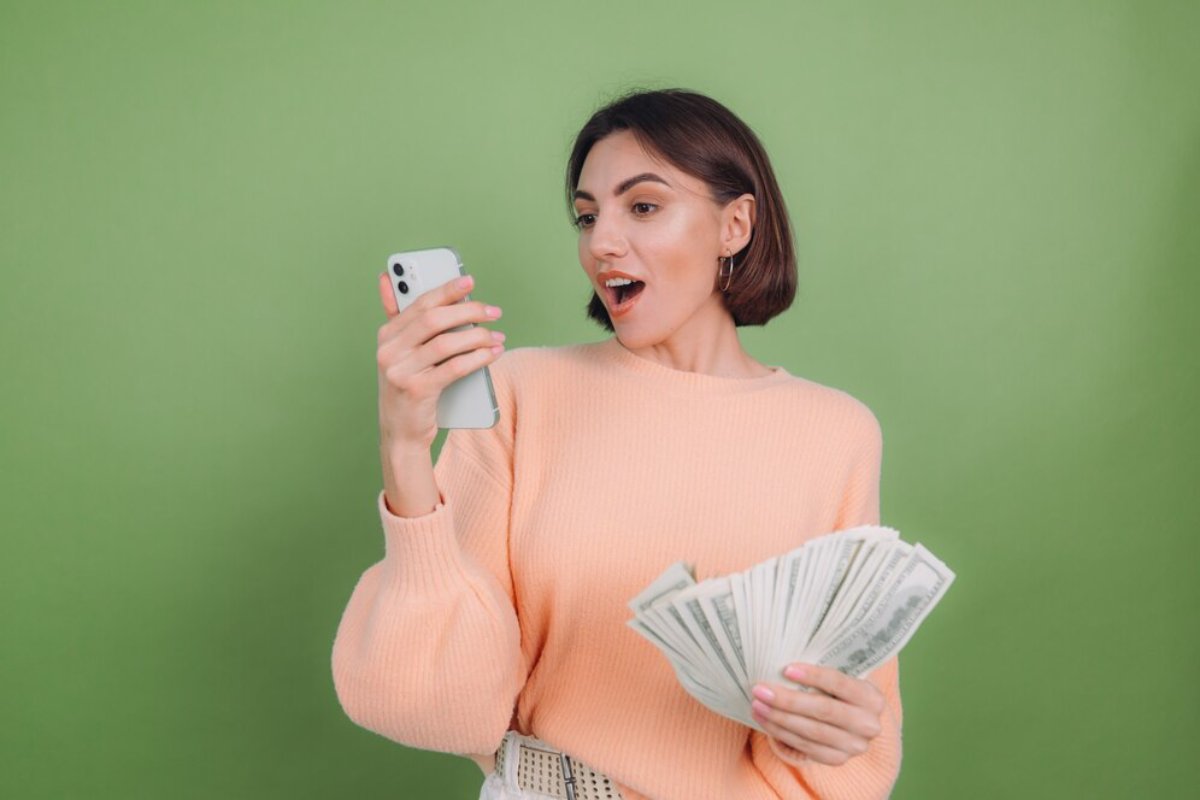 Woman in peach sweater holding cash and smartphone, looking surprised, against a green background.