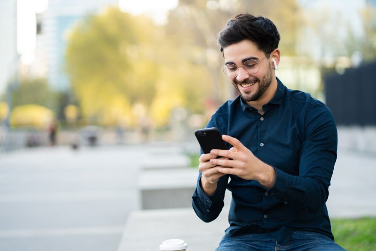 A joyful young man wearing glasses uses his smartphone outdoors, smiling as he reads the screen.