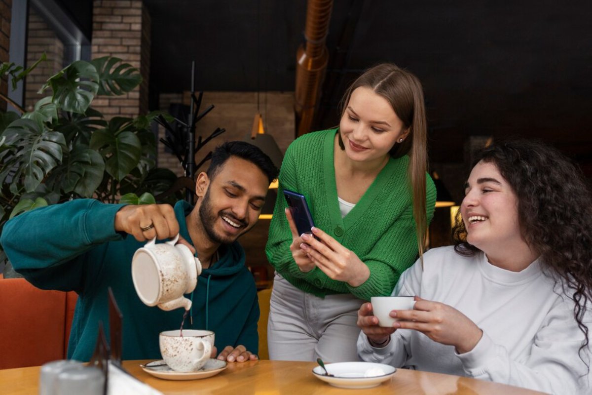 Three friends enjoy a lively moment in a cafe as one pours tea and another captures it on a smartphone.