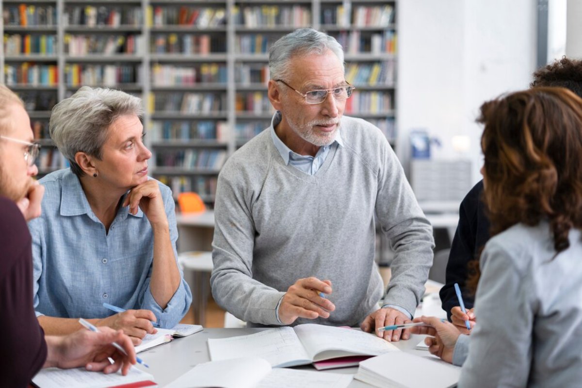 Senior adults and a young person in a discussion at a library table, surrounded by books.