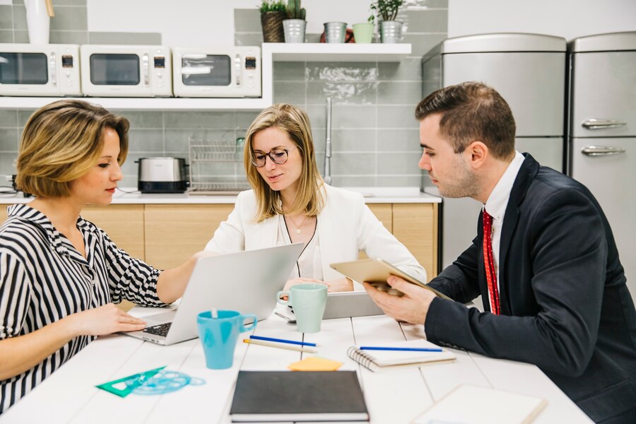 Three professionals collaborating around a laptop in a modern office kitchen.