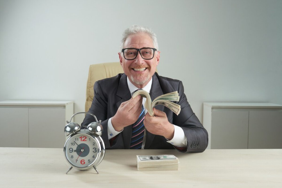 Happy older businessman with glasses counting money at a desk with a large alarm clock and stack of cash.