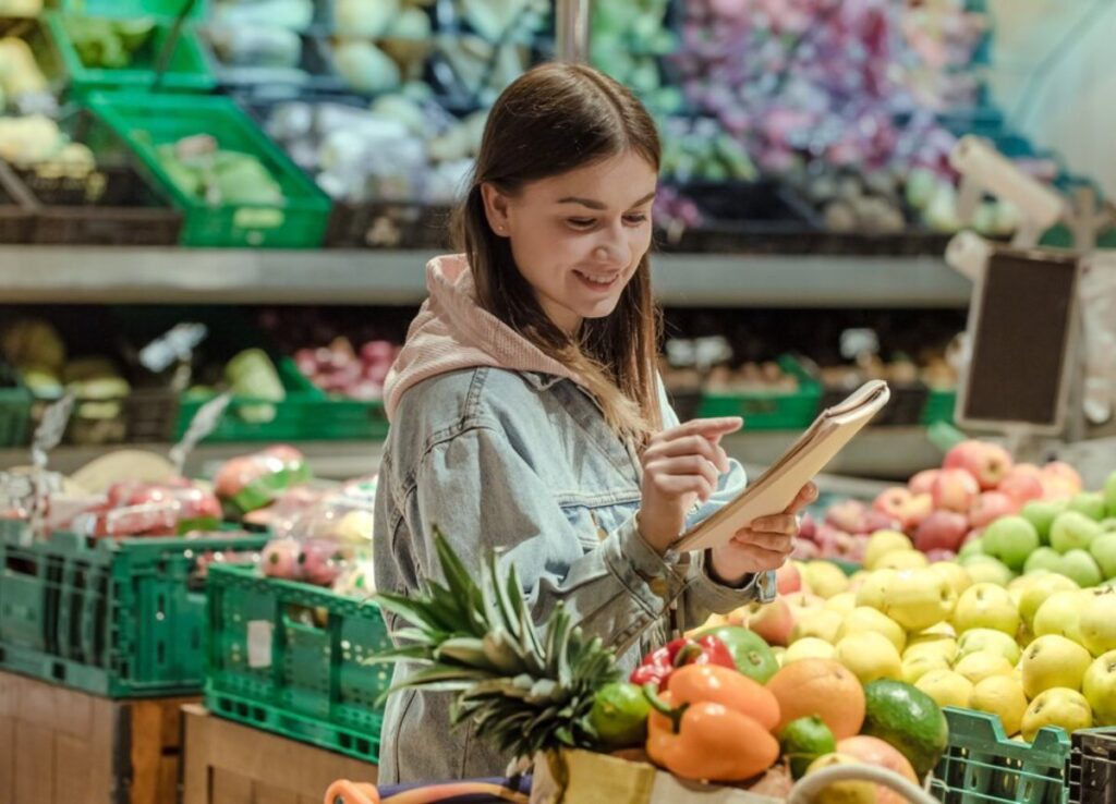A woman smiling and checking a tablet while shopping in a fruit section of a grocery store.