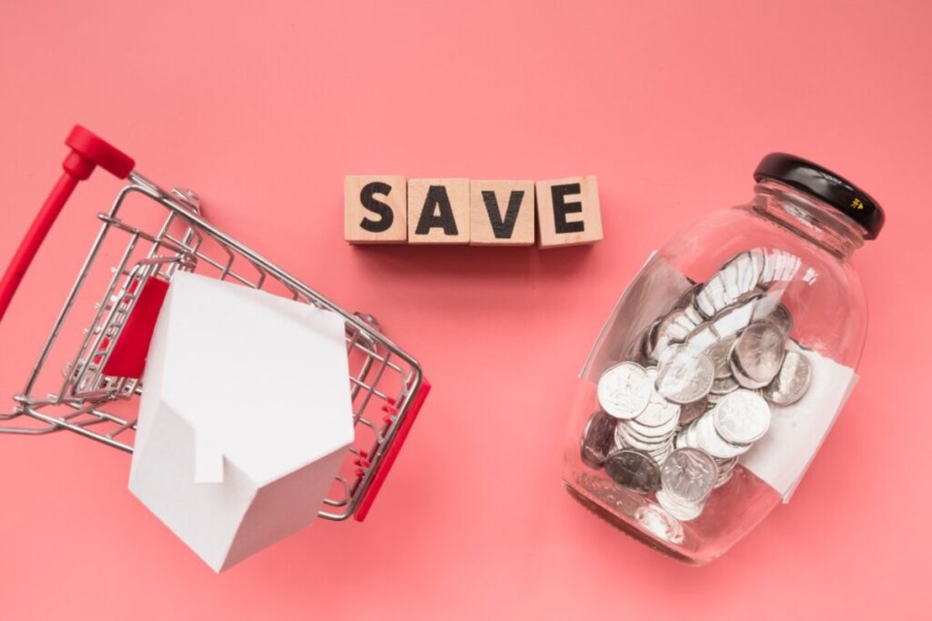 A miniature shopping cart with a house model, wooden blocks spelling "SAVE," and a jar of coins on a pink background.