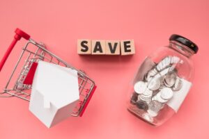 A miniature shopping cart with a house model, wooden blocks spelling "SAVE," and a jar of coins on a pink background.