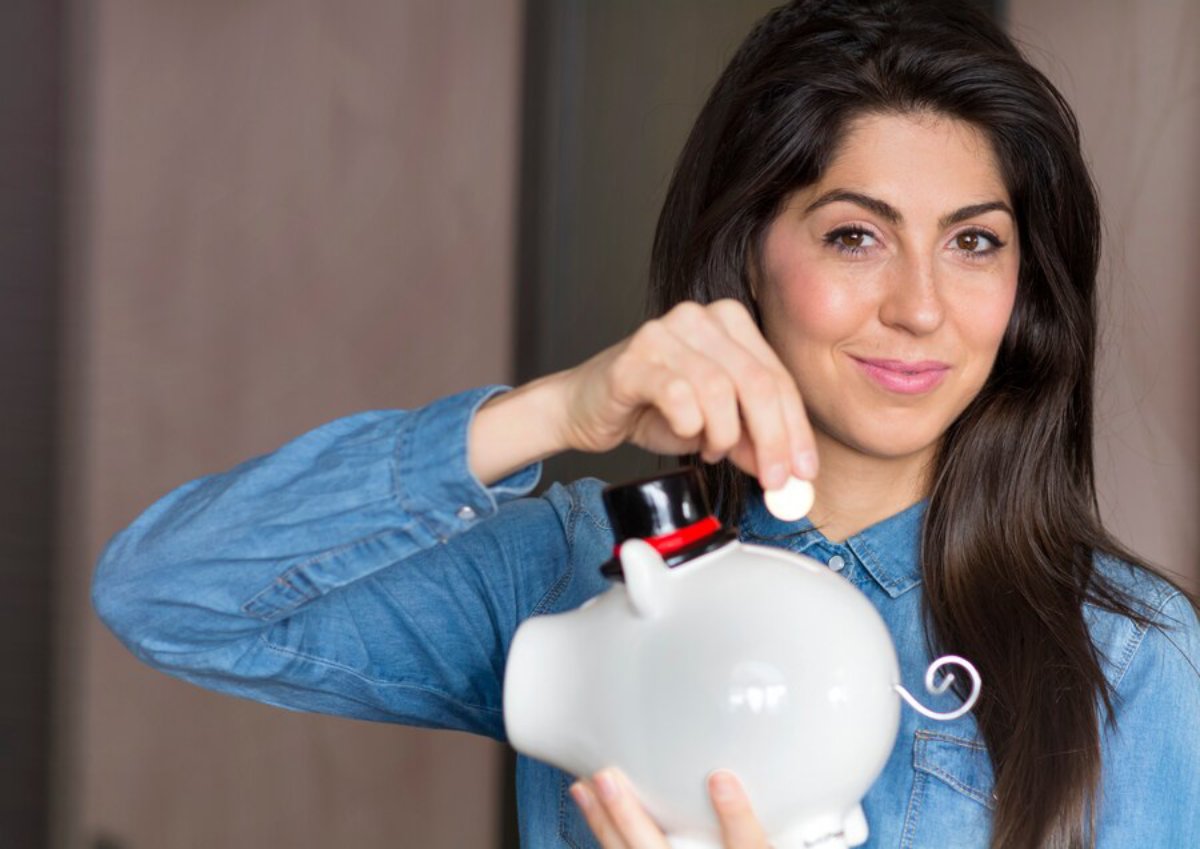 A woman in a denim shirt inserting a coin into a white piggy bank, smiling at the camera.