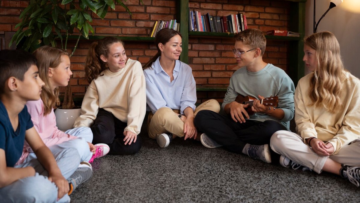 A teacher and diverse students sit on a carpet, engaging in a discussion while a boy plays a ukulele.