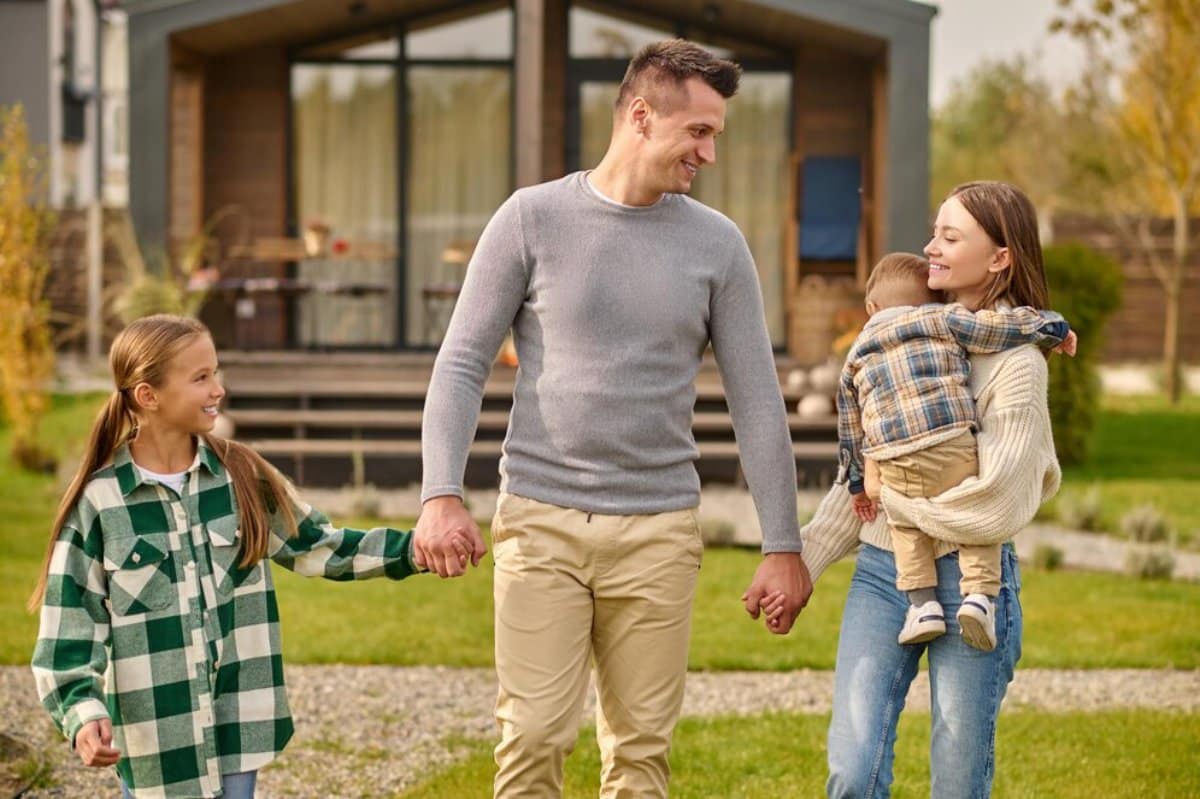 A father walks with his daughter and holds his son, smiling, in front of a modern house.