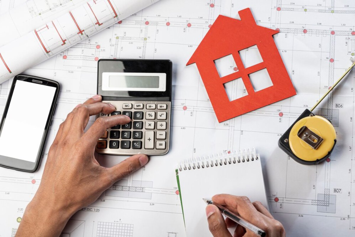 Hands calculating with a calculator and taking notes on architectural plans, with a house model and measuring tape nearby.