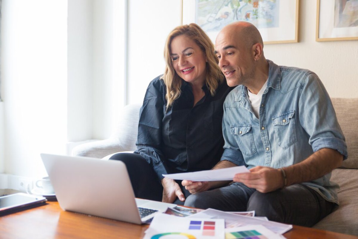A couple reviews documents together with a laptop on a couch in a bright living room.
