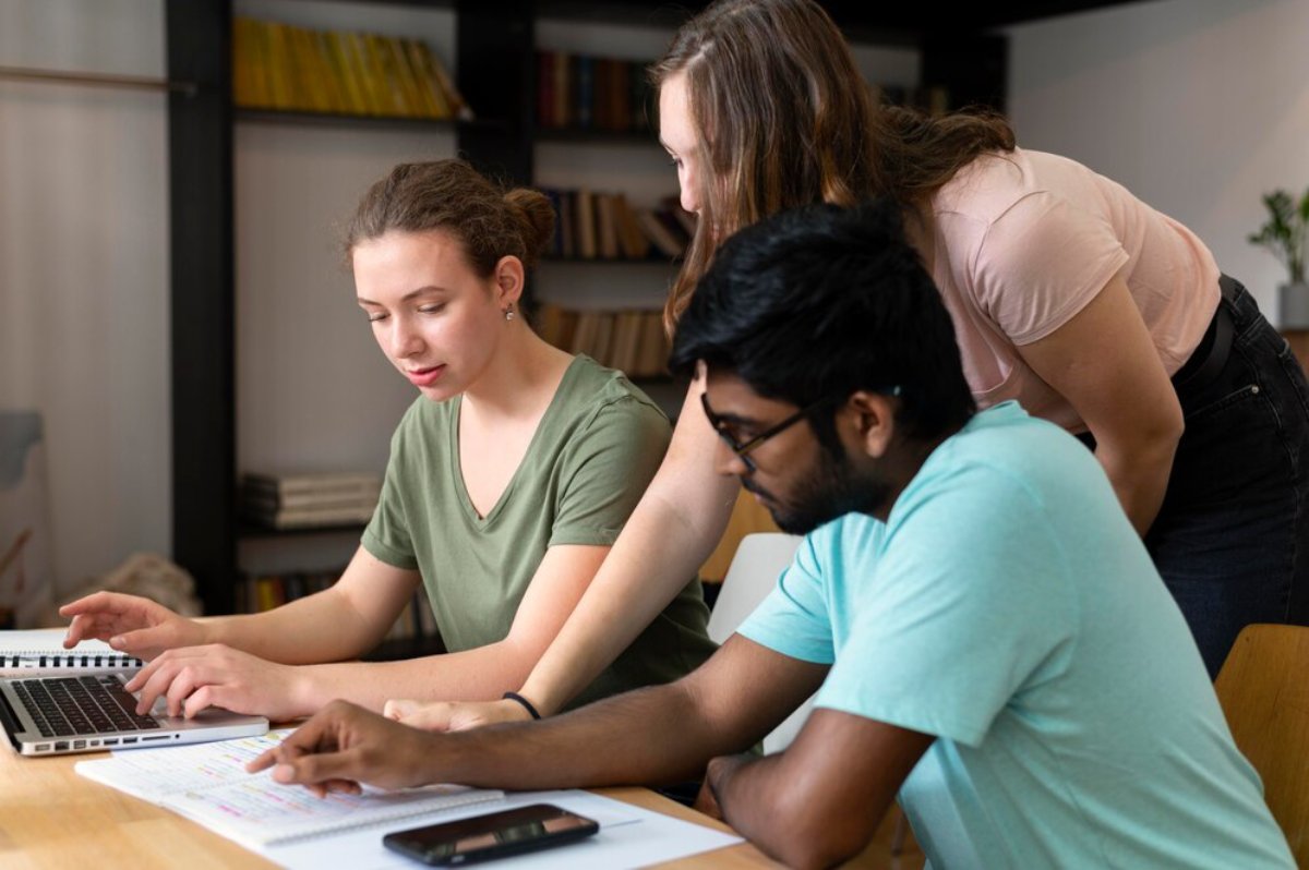 Three students collaborate on a project using a laptop at a wooden table in a library.