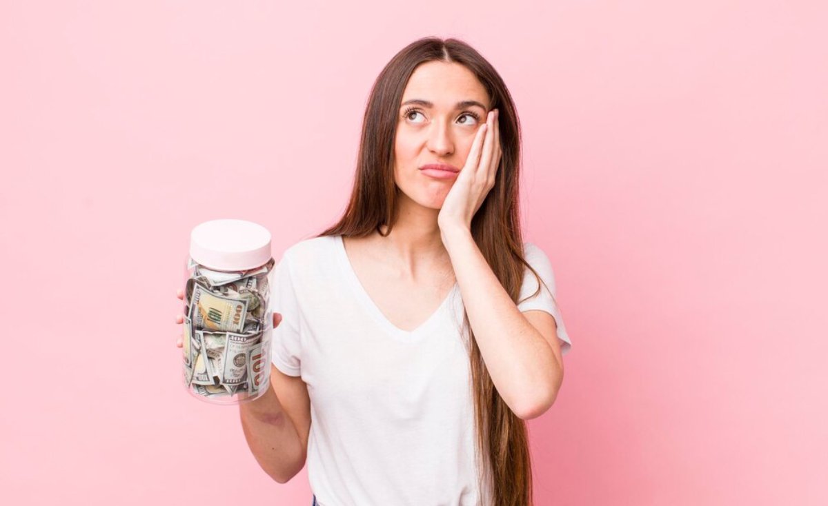 Woman in white T-shirt holding a jar of money, looking worried, against a pink background.