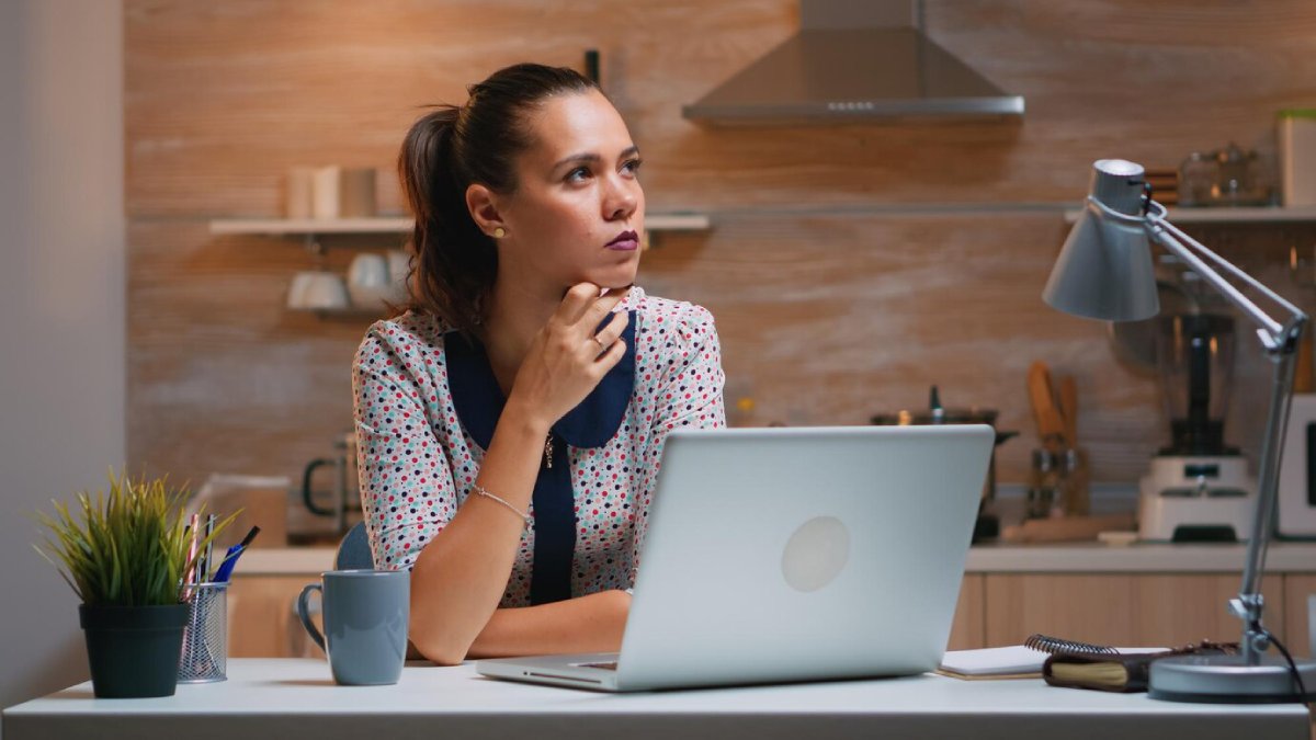 Woman with a thoughtful expression working at a home office desk with a laptop and desk lamp.