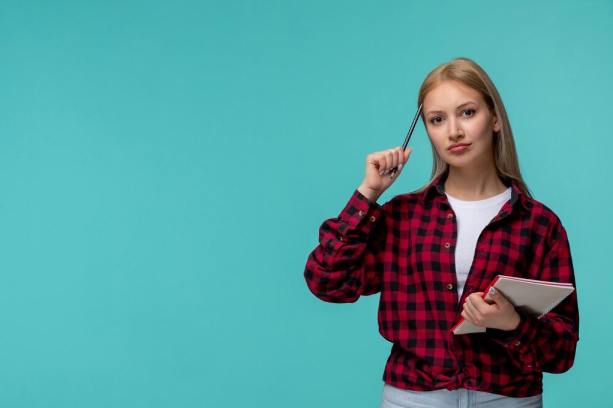Woman in red plaid shirt holding a pen to her temple, contemplating, with a notebook in her other hand, on turquoise background.
