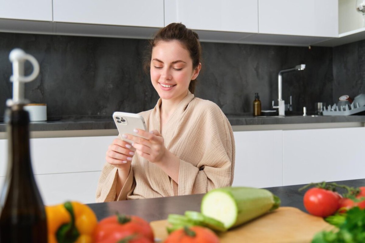 Woman in bathrobe smiling at phone in modern kitchen with vegetables on counter.