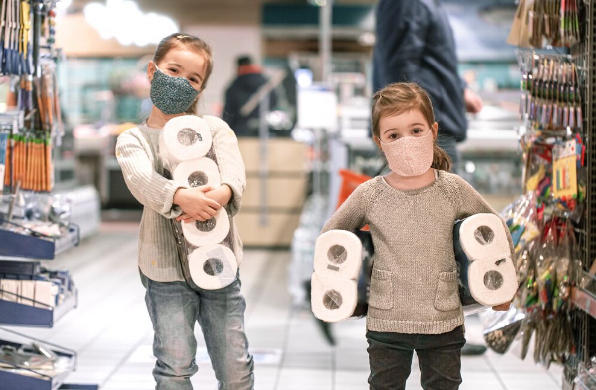 Two young children wearing masks and holding toilet paper packs in a supermarket aisle.