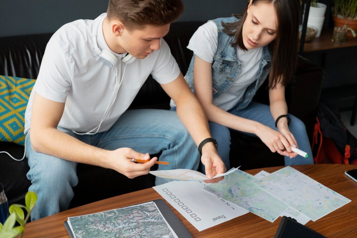 Two young adults examining and marking maps on a table, planning a trip or project together.