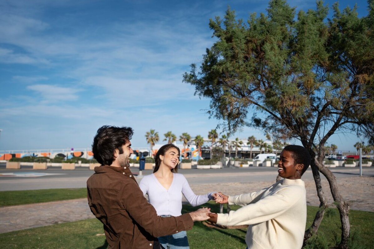 Three friends laugh and hold hands at a sunny coastal park with a tree and beachside shops in the background.