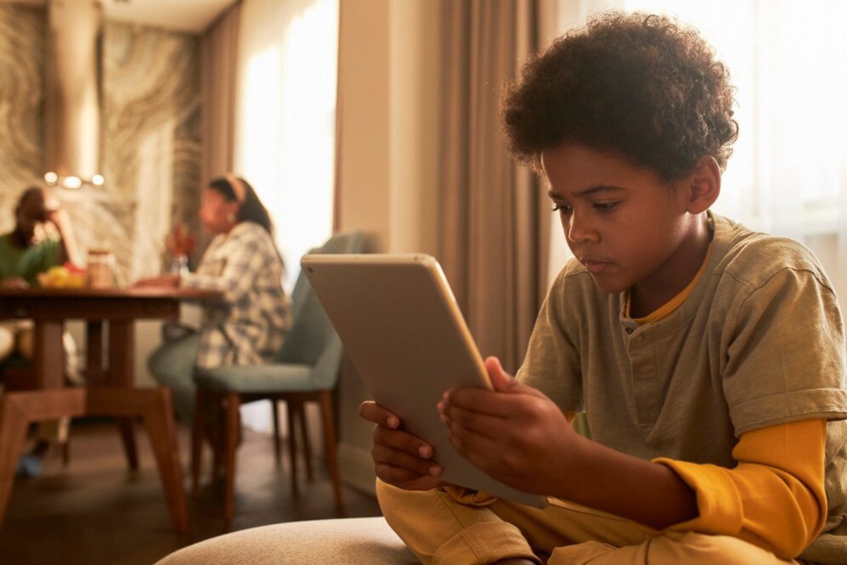 Young boy focused on using a tablet while his family converses in the background of a home dining room.