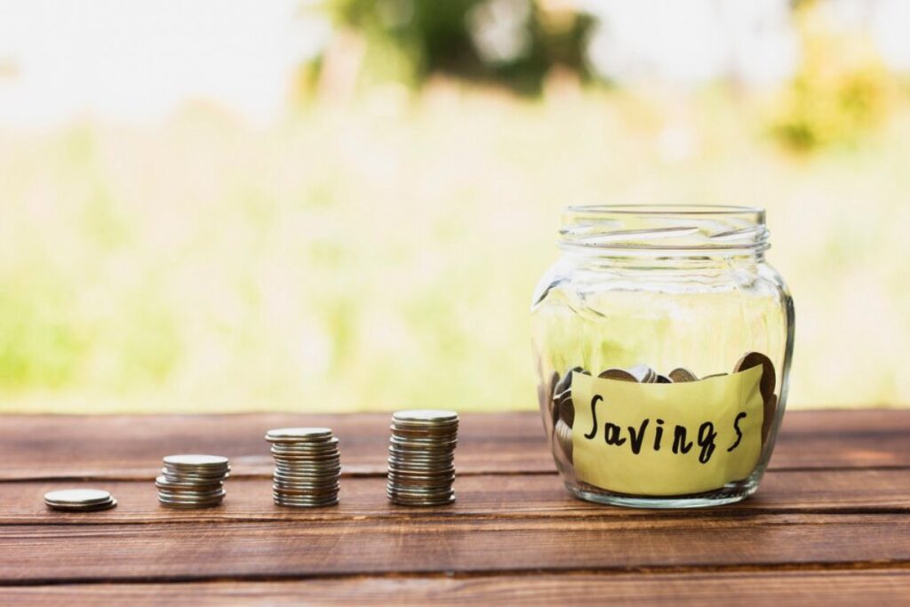 Coins stacked beside a glass jar labeled "Savings" on a wooden surface, outdoor blurred green background.