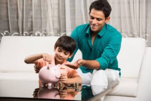 A man encouraging a young boy as he places coins into a piggy bank on a glass table indoors.