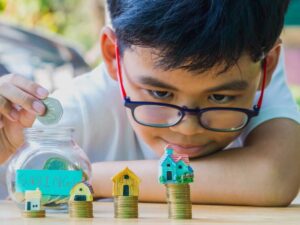 A child drops a coin into a jar labeled "savings," surrounded by stacks of coins and small house models, symbolizing financial growth.
