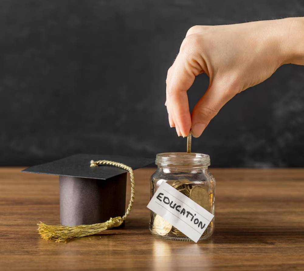 A hand placing a coin into a jar labeled EDUCATION beside a graduation cap on a wooden surface.