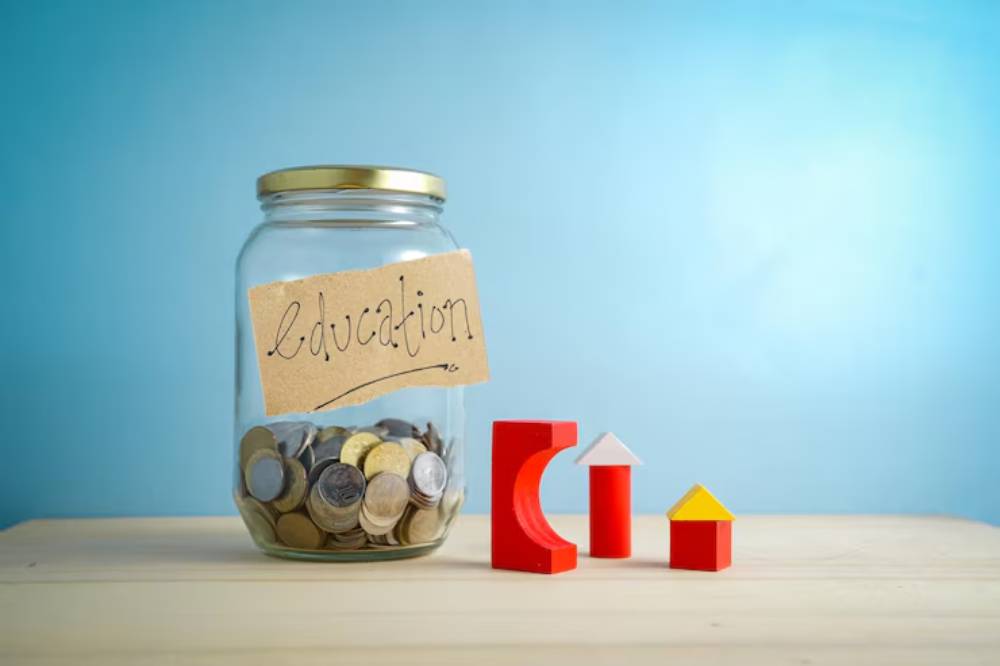 A glass jar labeled education filled with coins, next to colorful wooden building blocks on a pale wooden surface.