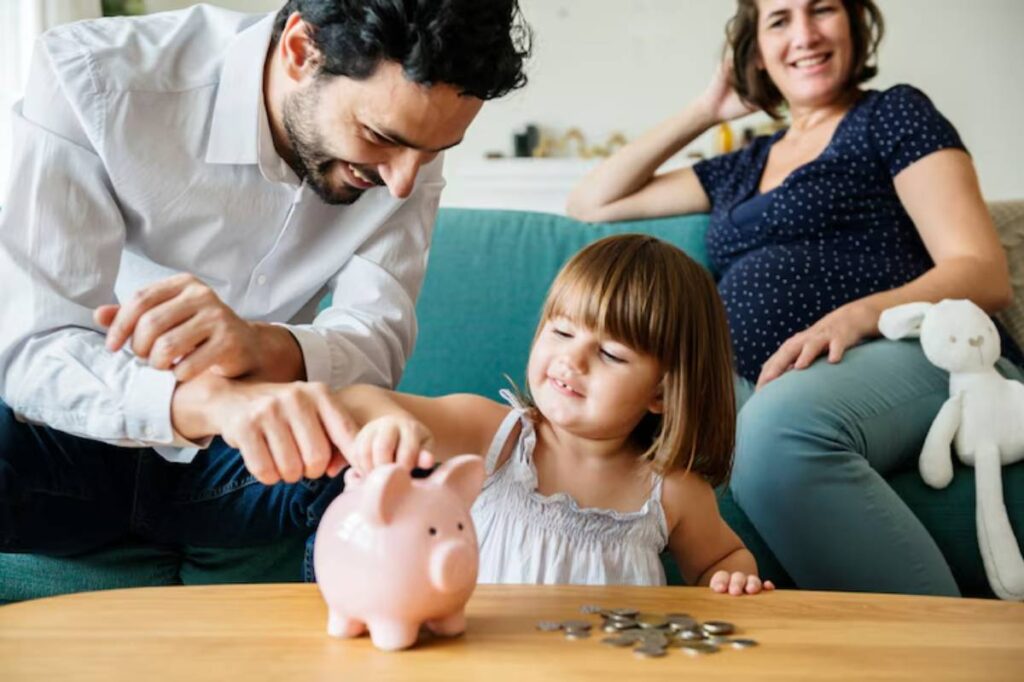 A child inserts coins into a pink piggy bank while an adult assists them, with a woman seated nearby on a couch.
