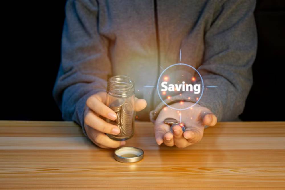 A person holding a jar of coins in one hand and a few coins in the other, with a glowing “Saving” label above.