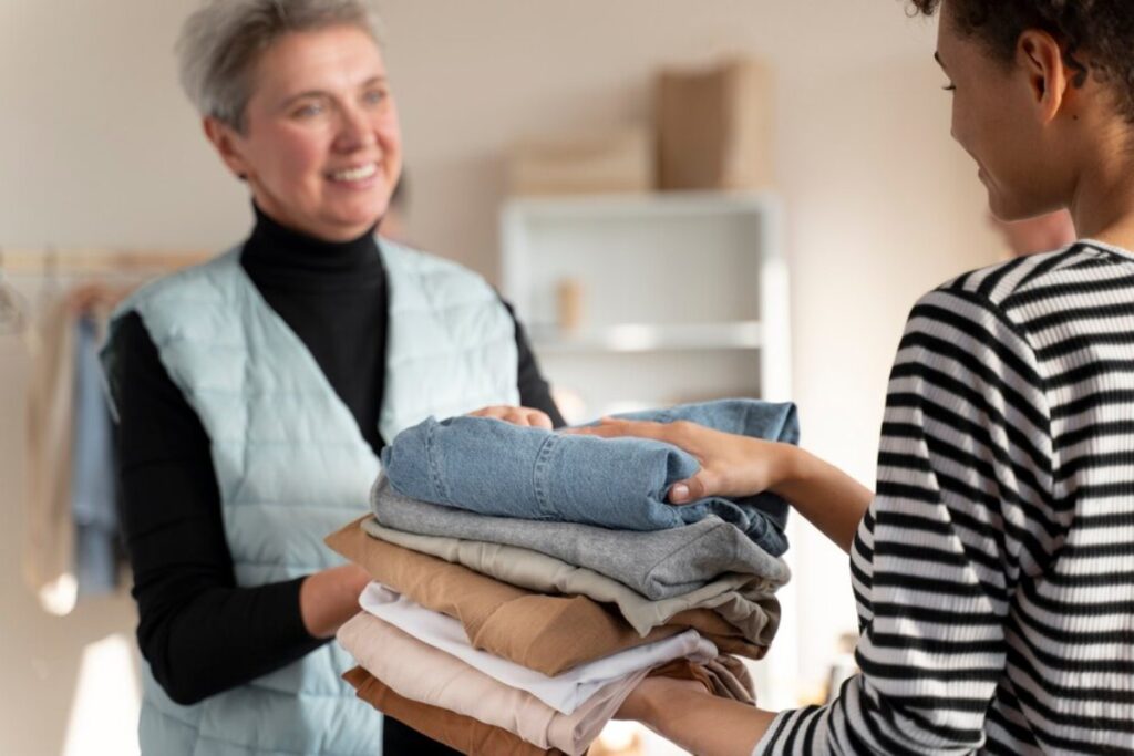 Two women exchanging a stack of folded clothes, one is smiling at the other in a room.