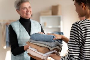 Two women exchanging a stack of folded clothes, one is smiling at the other in a room.